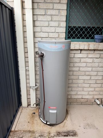 Old electric storage hot water tank mounted against a brick wall at a Gold Coast home before replacement.